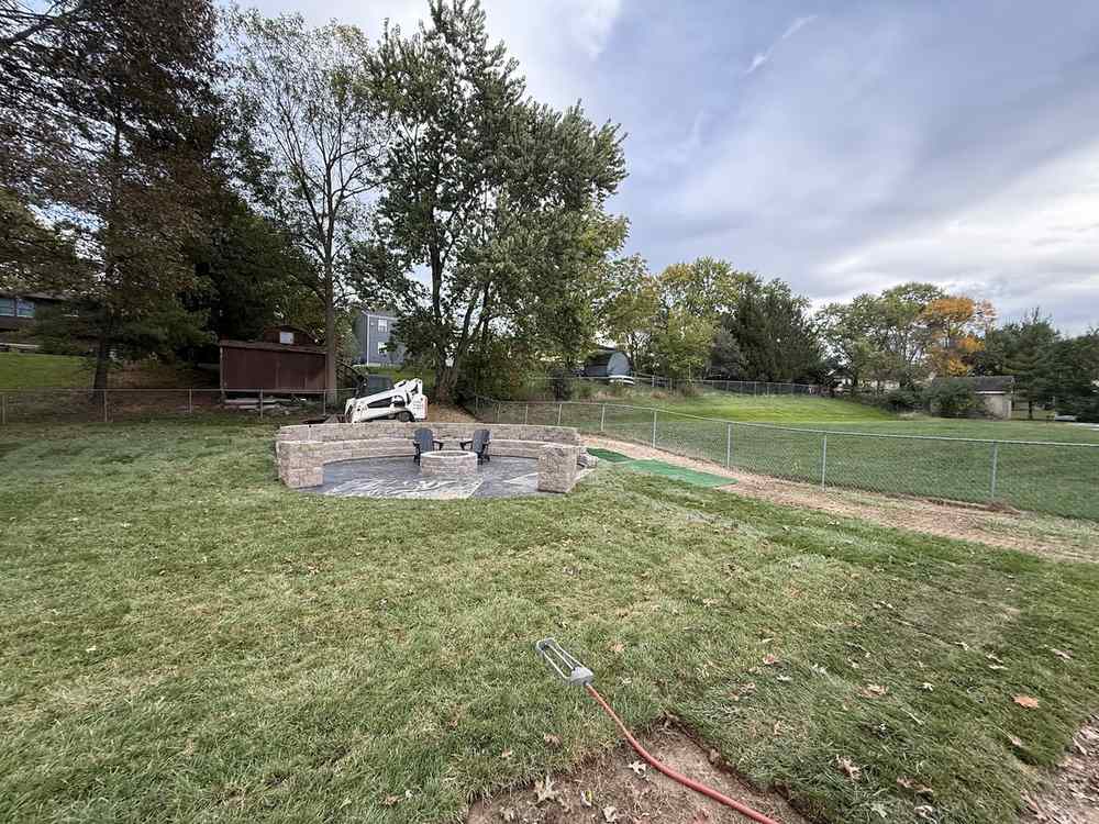 The same back yard with sod laid, a finished fire-pit patio with a seat wall, and a skid steer parked beside it.