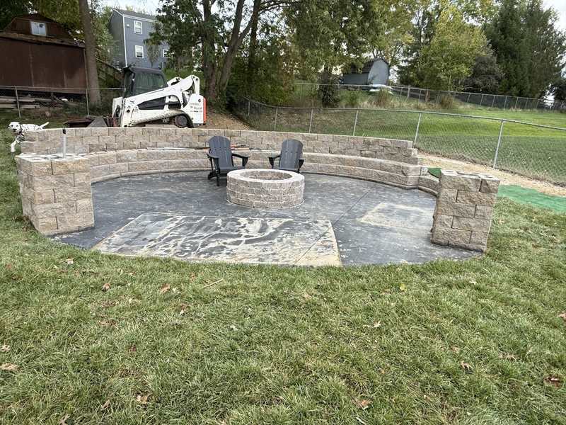 A finished circular patio with a block seat wall, a stone fire pit and two dark chairs, with new sod laid around it and a skid steer parked behind.